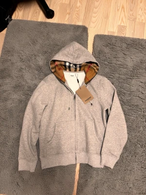 Grå burberry zip hoodie -  grå zip-hoodie från Burberry köpt för cirka 1 månad sen då den är rätt så liten på mig. Koftan är i mycket bra skick då den är köpt rätt så nyligen. 