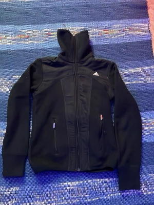 Svart ziptröja från Adidas - Säljer en svart ziptröja från Adidas med hög krage och två dragkedjeförsedda fickor framtill. Tröjan har klassiska vita Adidas-ränder på ryggen och liten logga på bröstet. Tillverkad i ett mjukt och stretchigt material, perfekt för en sportig look.