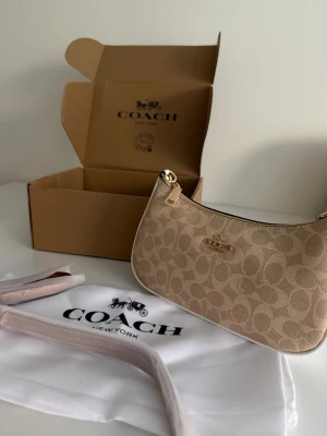 Beige axelväska från Coach - Snygg beige axelväska från Coach med klassiskt monogrammönster och guldfärgad logga framtill. Väskan har en stilren, avlång form och är tillverkad i canvas med detaljer i läder. Perfekt för dig som gillar trendiga accessoarer med lyxig känsla.