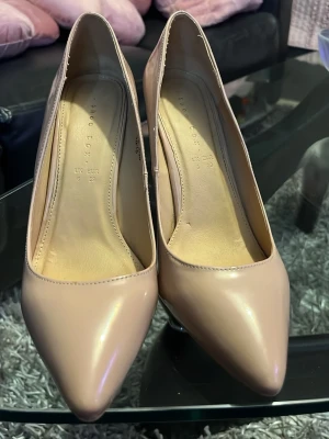 Beige lackpumps med spetsig tå - Snygga beige pumps med spetsig tå och hög klack i glansigt lackmaterial. Klassisk och stilren design som passar till många olika outfits. Skorna har en elegant siluett och är perfekta för dig som gillar en chic look.