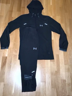 Svart Under Armour tracksuit - Säljer ett svart träningsset från Under Armour med huva och dragkedja på jackan samt matchande byxor. Setet har reflekterande detaljer och logga på både jacka och byxor. Perfekt för löpning eller gym. Materialet är lätt och andas.