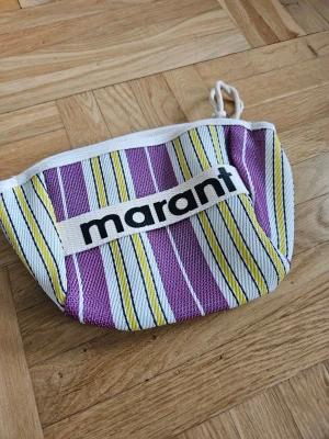 Randig necessär från Isabel Marant - Snygg necessär från Isabel Marant i tyg med lila, gula och vita ränder. Väskan har dragkedja upptill och ett tydligt Marant-tryck på framsidan. Perfekt för småprylar eller smink. Mjuk form och praktisk storlek.