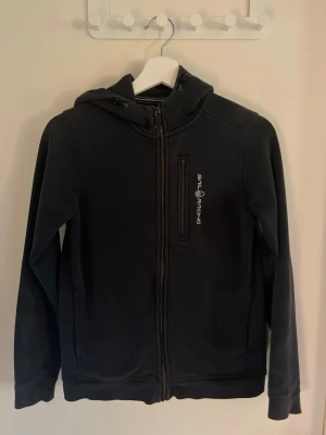 Svart hoodie från Sail Racing - Svart hoodie från Sail Racing den har lite solbränna med dragkedja framtill och logga på bröstet. Har en praktisk huva och en bröstficka med dragkedja dragkedjan är tyvär sönder. Tillverkad i mjukt material som passar perfekt till en avslappnad stil.