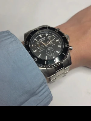 Champion Klocka - En klassisk Champion klocka med svart chronograf som har stenar runtom, urtavlan är 47mm bred och armlänken är ganska liten så den passar nog en smal handled men det medföljer 2 extra länkar som man kan fästa på för en större storlek