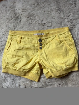 Gula shorts med knappar fram - Snygga gula shorts med tre knappar framtill och ribbade muddar i midja och benslut. Shortsen har fickor både fram och bak samt bälteshällor. Perfekta för soliga dagar och ger en fräsch look.