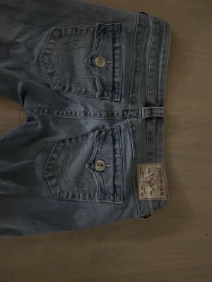 True Religion ljusblå bootcut jeans - Ljusblå jeans från True Religion med bootcut-passform och slitningar framtill. Klassiska bakfickor med dekorativa sömmar och silverfärgade knappar. Jeans i denim med patch på bakfickan.