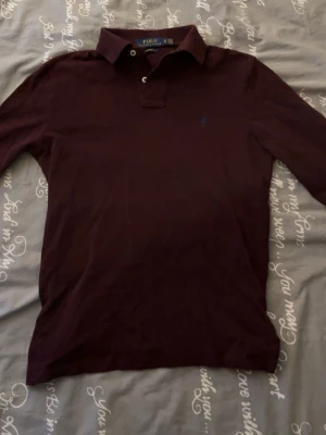Vinröd långärmad piké  Polo Ralph Lauren - Snygg vinröd långärmad skjorta från Polo Ralph Lauren med klassisk krage och diskret broderad logga på bröstet. Skjortan är i bomull och har en stilren look som passar perfekt till många olika outfits.