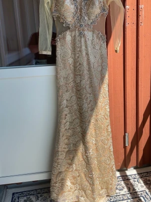 Champagnefärgad långklänning med spets - Elegant långklänning i champagnefärg med v-ringning, transparenta långärmar och detaljerad spets samt glittriga paljetter. Klänningen har markerad midja och vackra broderier över hela plagget. Perfekt för bal eller festliga tillfällen.