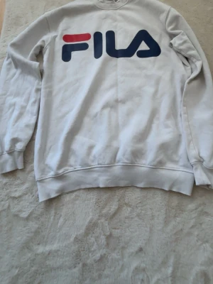 Vit sweatshirt från FILA - Klassisk vit sweatshirt från FILA med stor logga i blått och rött framtill. Tröjan har rund halsringning, långa ärmar och ribbade muddar vid ärmslut och nederkant. Perfekt för en sportig och avslappnad stil.