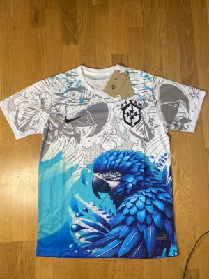 Brazil X Nike Dri fit shirt  - Unik sporttröja från Nike och Brasilien CBF med färgstark blå papegoja och grafiskt mönster i vitt, grått och blått. Tillverkad i Dri-Fit material som andas. Perfekt för dig som vill sticka ut på planen.