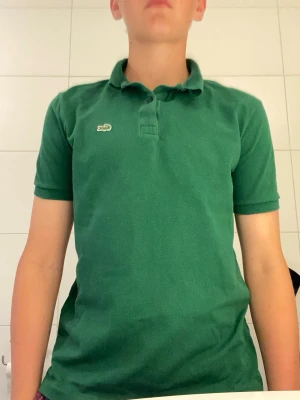 Grön pikétröja från Lacoste - Snygg grön pikétröja från Lacoste med klassisk krage och korta ärmar. Tröjan har den ikoniska krokodilloggan på bröstet och är tillverkad i mjuk bomull. Perfekt för en avslappnad och stilren look. Den börjar bli lite tajt så därför säljer jag den. Pris går att diskutera 