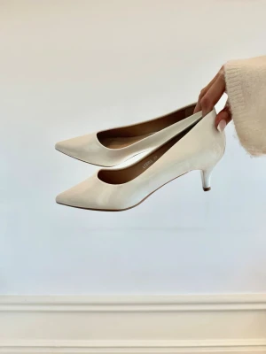 Vita klassiska klackar med låg klack - Säljer ett par stilrena vita klackar med spetsig tå och låg klack. Skorna har en elegant och tidlös design, perfekta för att lyfta vilken outfit som helst. Tillverkade i ett slätt material med diskret glans. 