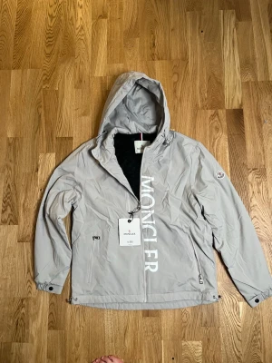 Ljusgrå vindjacka från Moncler - Snygg ljusgrå vindjacka från Moncler med huva och dragkedja framtill. Jackan har stora vita Moncler-bokstäver längs ena sidan, två dragkedjeförsedda fickor och justerbar nederkant. Materialet är tunt och vattenavvisande, perfekt för blåsiga dagar.HELT NY HAR I SVART OCKSÅ