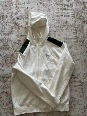 Vit hoodie med zip från Nike - Vit hoodie från Nike med hel dragkedja och huva. Svarta detaljer på axlarna och klassisk Nike-logga på bröstet. Mjuk bomullsmix och praktiska fickor framtill. Perfekt för en avslappnad och sportig look.