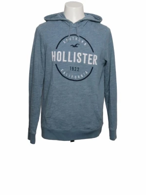 Blå hoodie från Hollister - Snygg blå hoodie från Hollister med stor logga och texten 'Southern California 1922' på bröstet. Hoodien har huva med dragsko, magficka och ribbade muddar vid ärmslut och nederkant. Perfekt för en avslappnad och cool stil.