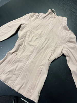 Ljusrosa träningsjacka från Lululemon - Snygg ljusrosa träningsjacka från Lululemon med figurnära passform, hög krage och hel dragkedja framtill. Jackan har två fickor med dragkedja och är tillverkad i ett stretchigt, mjukt material som passar perfekt till träning eller promenader.