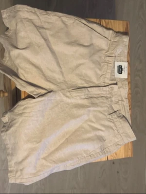 Beige linneshorts från COS - Snygga beige shorts från COS i luftig linnemix. Klassisk design med fickor fram och bak, bälteshällor och knappstängning. Perfekta för varma dagar och enkel att matcha med olika toppar.