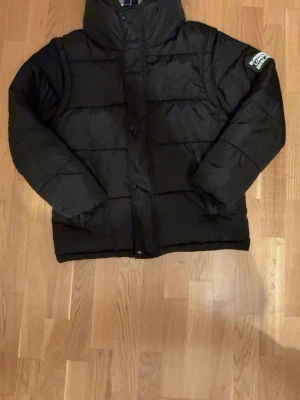 Svart pufferjacka Burberry - Säljer en svart pufferjacka från burrbery med avtagbara ärmar så den kan användas som väst. Jackan har hög krage, dragkedja och tryckknappar samt två sidofickor med dragkedja. Insidan av kragen har rutigt foder.