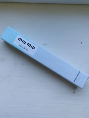 Miu Miu Fleur de Lait Eau de Parfum - Miu Miu Fleur de Lait Eau de Parfum i en elegant, avlång förpackning i ljusblått och vitt. Märkets logga syns tydligt på framsidan. Perfekt för dig som gillar fräscha och blommiga dofter med en lyxig touch.