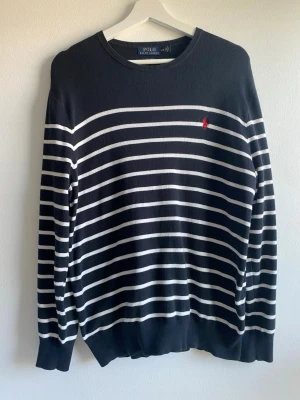 Randig tröja från Polo Ralph Lauren M - Tröja från Polo Ralph Lauren med vita ränder och röd broderad logga på bröstet. Tröjan har rund halsringning och är tillverkad i mjukt bomullsmaterial. Perfekt för en stilren och klassisk look.
