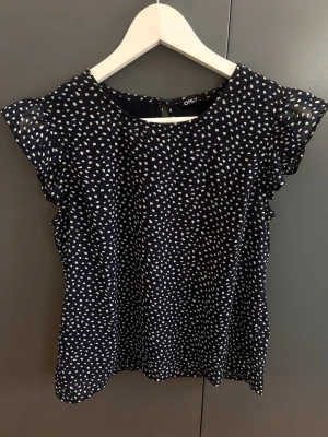 Mörkblå prickig blus från ONLY - Snygg mörkblå blus från ONLY med vita prickar och volangdetaljer vid ärmarna. Passar också S! Blusen har rund halsringning och är tillverkad i ett lätt, luftigt tyg som känns skönt mot huden. Perfekt för en trendig och avslappnad look. 