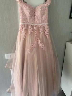 Lång ljusrosa spetsklänning - Romantisk långklänning i ljusrosa med vackra spetsdetaljer och blommiga applikationer. Klänningen har smala axelband och ett lätt, svepande tyg som ger en drömmig känsla. Perfekt för speciella tillfällen när du vill känna dig extra fin. Köpt för 2200