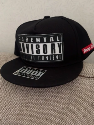 Svart snapback Parental Advisory Keps - Svart snapback-keps från Damp Lush med stort 'Parental Advisory Explicit Content'-tryck framtill och mindre patch på skärmen. Röd broderad logga på sidan. Klassisk platt skärm och justerbar baksida. Perfekt för en streetwear-look.