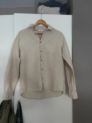 Beige linneskjorta från Lindex - Säljer en stilren beige skjorta från Lindex i linne. Skjortan har klassisk krage, bröstficka och knappar framtill. Långärmad modell med avslappnad passform, storlek 170.