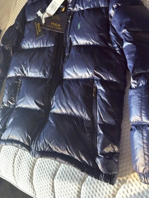 NY! Polo Ralph Lauren jacka - Snygg mörkblå pufferjacka från Polo Ralph Lauren med glansig finish och klassisk grön logga på bröstet. Jackan har dragkedjor på fickorna och en hög krage. Perfekt för kalla vinterdagar och riktigt trendig stil.