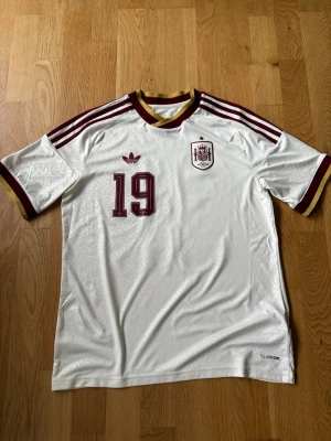 Vit Lamine Yamal Spanien matchtröja - Tröjan är helt ny och oanvänd då jag köpte en för liten storlek. Tröjan är köpt från Unisport i Stockholm för 1149 kr. Skriv gärna vid funderingar.               Säljer en vit matchtröja från Adidas med Spaniens landslagsemblem och nummer 19 i vinrött. Tröjan har vinröda och gula detaljer på krage och ärmslut samt klassiska Adidas-ränder på axlarna. Tillverkad i lätt och ventilerande material, perfekt för fotboll.      