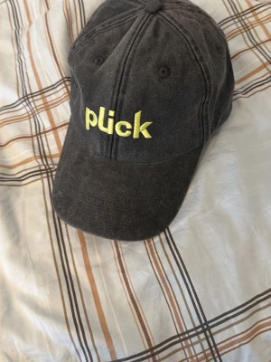 Svart keps med gul brodyr från Plick - Säljer en svart keps från Plick med gul broderad text framtill. Klassisk böjd skärm och justerbart spänne i metall baktill. Tillverkad i bomullstyg och passar perfekt till en avslappnad streetwear-look.
