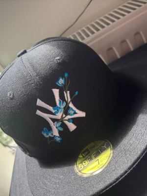 Svart NY Yankees keps med blommor - Snygg svart New Era 59FIFTY keps med NY Yankees-logga framtill i vitt och blå broderade blommor. På sidan finns ett blått World Series-märke. Klassisk flat peak och broderade detaljer. Perfekt för dig som vill sticka ut med din stil.