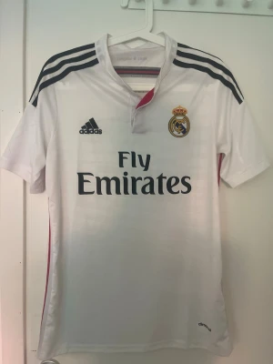 Real Madrid 14/15 tröja - Snygg vit Real Madrid matchtröja från Adidas med svarta ränder på axlarna och klubbmärke på bröstet. Tryck med Fly Emirates på framsidan och Climacool-material som andas. Perfekt för fotbollsträning eller att bära till vardags. Kom privat vid funderingar! 