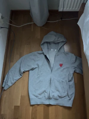 Grå hoodie Comme des Garçons Play - Säljer en grå hoodie från Comme des Garçons Play. Den är i bra skick men har en liten defekt vad jag har sett bara. Det är dragkedjan som har gåt av lite, själva ”handtaget” för zippern. Är inget som påverkar funktionen på det sättet dock. Den är M i storleken men skulle säga den sitter rätt fint på S.                                      Priset kan självklart diskuteras.                                                  Vid frågor är de bara och höra av sig