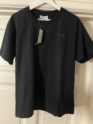 Svart t-shirt från Lacoste - En stilren svart t-shirt från Lacoste med diskret logga på bröstet. Klassisk rund halsringning och korta ärmar. Tillverkad i mjuk bomull för en bekväm känsla. Perfekt basplagg för en enkel och snygg look.