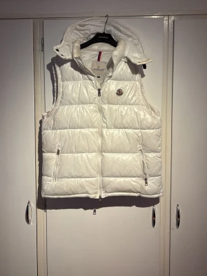Vit dunväst från Moncler med huva - Snygg vit dunväst från Moncler med avtagbar huva och två dragkedjeförsedda fickor. Västen har quiltad design och Moncler-logga på bröstet. Perfekt för lager-på-lager och håller dig varm med stil. Klassisk passform och tidlös look.
