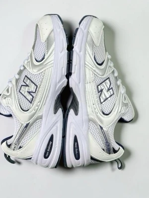 Vita New Balance 530 sneakers - Fräscha vita New Balance 530 sneakers med silvriga och grå detaljer. Skorna har meshpaneler för extra andningsförmåga och klassisk logga på sidan. Snygg chunky sula och sportig vibe, perfekta för en trendig streetstyle-look.