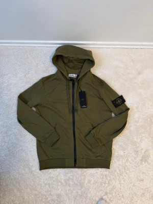 Olivgrön hoodie från Stone Island - Säljer en olivgrön hoodie från Stone Island med dragkedja framtill och klassisk patch på vänster ärm. Hoodien har huva med dragsko, ribbade muddar och känguruficka. Perfekt för dig som gillar streetwear och vill ha en snygg och bekväm look.