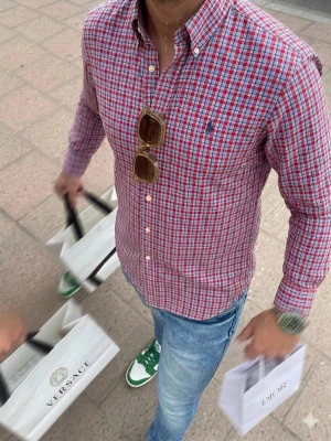 Rutig skjorta från Ralph Lauren - Riktigt snygg rutig skjorta från Ralph Lauren i rött, blått och vitt. Klassisk button-down krage, långa ärmar och broderad logga på bröstet. Perfekt till jeans eller chinos för en clean och stilren look. Har ett litet hål. Se bild 3