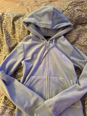 Ljusblå velour hoodie Juicy Couture - Supermjuk ljusblå hoodie i velour från Juicy Couture. Dragkedja framtill, stor huva och känguruficka. Ikonisk JC-logga broderad på bröstet. Perfekt för en chill och trendig look.