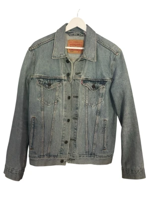 Levi's klassisk blå jeansjacka - Snygg blå jeansjacka från Levi's med raka linjer, två bröstfickor med lock och knappar framtill. Jackan har klassisk krage, knappstängning och detaljerade sömmar. Tillverkad i slitstarkt denimtyg som ger en tidlös look.