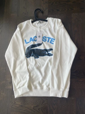 Vit sweatshirt från Lacoste - Helt ny och oanvänd Lacoste sweatshirt med tags kvar. Storlek M men liten i storleken, passar även S. Svart med stort Lacoste-tryck framtill i blått och vitt. Skön passform och perfekt till vardag/streetwear