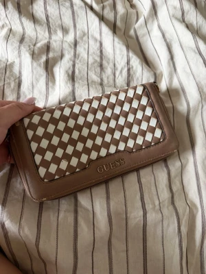 Beige och vit plånbok från Guess - Snygg plånbok från Guess i beige och vitt med flätad design på framsidan. Plånboken har dragkedja runtom, flera kortfack och ett myntfack med dragkedja. Tillverkad i konstläder och har Guess-loggan framtill. Fåtal skavanker som syns på bilden.