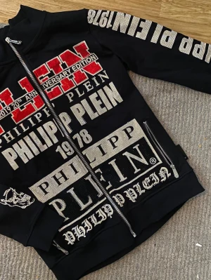 Philipp Plein hoodie med tryck - Svart hoodie från Philipp Plein med stora vita och röda tryck och logotyper över hela plagget. Dragkedja framtill och detaljer på ärmarna. Snygg streetwear-stil med markerad branding och coola detaljer.