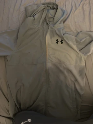 Ljusgrå hoodie från Under Armour - Snygg ljusgrå hoodie från Under Armour med dragkedja och svart logga på bröstet. Hoodien har huva och två fickor framtill. Perfekt för en sportig och avslappnad stil.