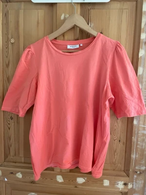 Korallrosa t-shirt från MSCH - Snygg korallrosa t-shirt från MSCH Copenhagen med rund halsringning och puffiga korta ärmar. Tillverkad i mjuk bomull, perfekt för en chill och trendig look. Enkel design som ändå sticker ut med sin färg och ärmdetalj.