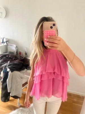 Rosa volangtop med lager på lager - Snygg rosa ärmlös topp med flera lager volanger och lös passform. Perfekt för dig som vill sticka ut med en färgglad och luftig stil. Toppen har en hög halsringning och är tillverkad i ett lätt, transparent material.