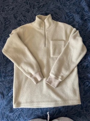 Beige teddytröja med halv dragkedja  - Mysig beige teddytröja från J&J med hög krage och halv dragkedja framtill. Tröjan har en bröstficka och ribbade muddar vid ärmsluten. Perfekt för kalla dagar och enkel att matcha med jeans eller mjukisbyxor. Passar S/M