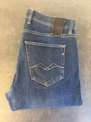 Blå jeans från Replay stl 34/32 - Snygga blå jeans från Replay med klassisk femficksmodell och raka ben. Jeansen har en cool bakficka med broderad detalj och läderpatch i midjan. Tillverkade i mjukt denimtyg som ger en skön passform och stilren look.