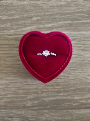 Silverfärgad ring med gnistrande sten - Snygg silverfärgad ring med en stor rund klar sten i mitten och mindre stenar på sidorna. Ringen har en elegant och klassisk design som verkligen fångar blicken. Levereras i en hjärtformad röd sammetsask.